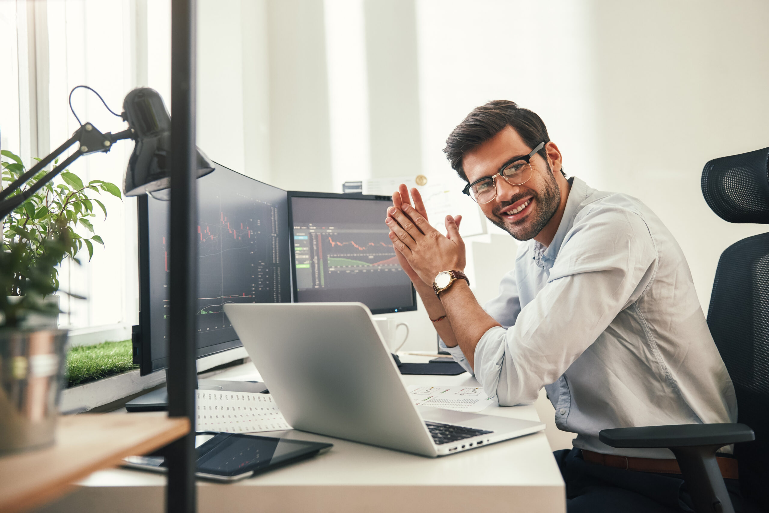 Young and successful. Happy bearded trader in formal wear and eyeglasses looking at camera and smiling while sitting in his modern office. Stock broker. Forex market. Trade concept